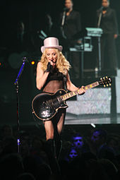 A blond woman standing on a stage. She has curvy, flowing hair and is dressed in a black, translucent top with boots in her leg and a white hat. The woman is holding an electric guitar with her left hand and singing in to a microphone in her right. She is surrounded by audience members whose heads can be seen in the image. Behind the woman, tow back-up singers can be seen in the distance.