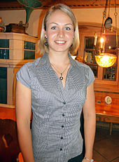 A blonde woman in casual clothes smilies into the camera. She wears a top with a narrow pattern of black and white strips and stands in a room with traditional Bavarian interior.