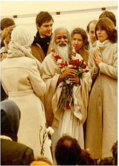 A photograph of the Maharishi holding flowers and surrounded by people wearing coats.