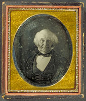 Half-length photographic portrait of an elderly, balding man dressed in a dark coat, vest and cravat