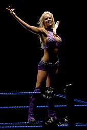 A blonde Caucasian woman posing on the turnbuckles in a wrestling ring with both her hands in the air. She is wearing a purple crop top, purple shorts with a silver belt and purple wrestling boots.