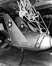 Black-and-white photograph of small egg-shaped fighter aircraft hoisted by a lifting mechanism underneath a large bomber.
