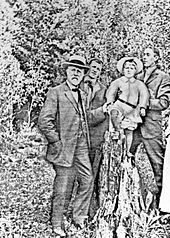 Faded, grainy image of three men in the outdoors, holding up a boy. The man on the left has a short white beard and mustache, a hat, and a three-piece suit.