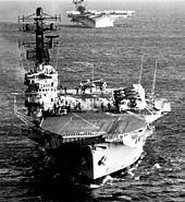 An aerial photo of an aircraft carrier with several aircraft on her flight deck. Another carrier is visible in the background.