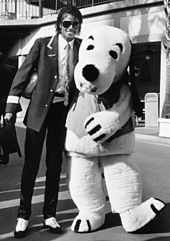 A black and white image shows a man standing next to a person dressed in a full dog costume. The man on the left has his left arm around the waist of the other person and is smiling.