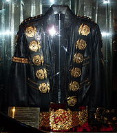 A black jacket with five round golden medals on its left and right shoulders and a gold band on its left arm sleeve. The jacket has two belt straps on the right bottom sleeve. Underneath the jacket is a golden belt, with a round ornament in its center. There is a red light reflecting on the jacket and belt as well as a gold colored plate on the left side of the jacket and belt.