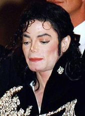 A close-up image of a pale skinned man with black hair. He is wearing a black jacket with white designs on it.