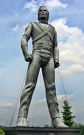 A silver colored statue of a male. The statue is placed standing up with its arms bent inward and both legs spaced apart. The statue's clothes have wrinkles and it is wearing heeled shoes. In the background, a tree and a light blue sky with multiple clouds can be seen.