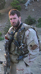 Michael Murphy in tan and brown desert camouflage looking at the camera. He is wearing several pieces of green military combat gear and is holding a weapon. There is a hill behind him covered in rocks, dirt and sticks.
