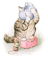 A cat standing on a red cushion with its head wrapped in a blue cloth peeks at a mouse in a green jacket