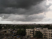 Daytime view over a city: de-laminating concrete housing blocks surrounded by rough slum tenements. In the middle distance, an expanse of trees: perhaps a park. Near the horizon, the largely concrete structures that compose the city continue. The whole is enveloped by an ominous sky filled with storm clouds promising imminent heavy rain.