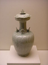 A white, handle-less jar with a small base, a wider body, and then a long, thin, opening at the top. Four flower-shaped ornaments are attached to the point where the body and the stem of the jug meet.