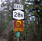 Four signs are mounted on a pole. From top to bottom, they are: the word "east", a NY 28N shield, a yellow-on-brown sign with an outline of Theodore Roosevelt's face and the text "Roosevelt-Marcy Trail", and a reference marker for NY 28N.