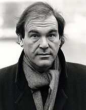 A man, wearing a coat and scarf, furrows his brow.