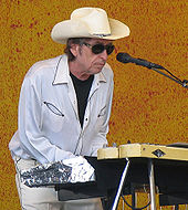 Dylan, wearing a white shirt and pants, sunglasses and a cowboy hat, plays the keyboards onstage.