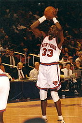 A basketball player attempting a jump shot. He is wearing a white jersey and shorts, both with orange stripes. The jersey has an orange "NEW YORK" and "33" on the front, and the shorts have a logo with an orange "KNICKS" above a basketball.