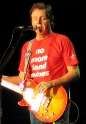 McCartney, in his late sixties, playing an orange electric guitar and wearing a red shirt that bears, in white writing, the words "no more land mines." His eyes are closed.