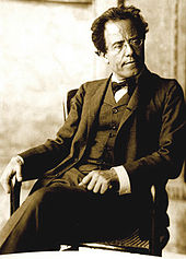  Middle-aged man, seated, facing towards the left with head turned towards the right. He has a high forehead and rimless glasses, and is wearing a dark, crumpled suit