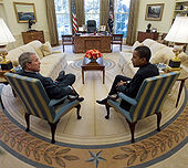 Obama meets with Bush in the Oval Office. Both sit at a distance in front of the presidential desk with their legs crossed and their backs on an angle toward the camera. They sit at right angles to each other.