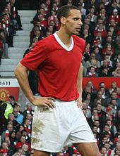A photograph of a man with very short, dark hair. He is standing with his hands on his hips, and he is wearing a plain red shirt with a white collar and white shorts.