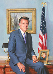 Mitt Romney resting on a wooden desk, flanked by an American flag, a picture of his wife, a lamp, and a painting of mountains