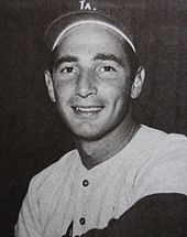 A black-and-white photograph of a smiling man in a baseball cap with an interlocking white "LA" on the front