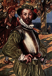 A bearded man, wearing green shirt and doublons, stands with his hands on his hips. The sea is visible in the background. A cuirass (metal armor) protects his upper torso.  He is also wearing a green hat with a red rose and a white ruff on his neck.  A rapier hangs ready at his left hip.  Behind him on his right, a man is walking by, carrying a laden rucksack over his shoulder.