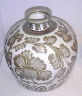 A handle-less jug which has a small bottom, a wide middle, and a very small opening at the top. The jug is white porcelain with a tan illustration of leaves or a vine wrapping around the entirety of the jug.