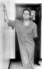 Bundy casually leans on the wall while dressed in prison garb.