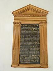 A photograph of a memorial in a wooden frame hanging on the wall of a church. The frame is a carved depiction of a Greek temple with columns and a small gable. The inscription is of gold lettering on a black background