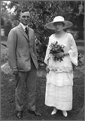 Wedding photo of man in gray suit and woman in hat with white dress holding flowers