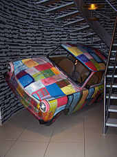 A car with bright coloured squares painted on the exterior is tilted slightly to its left side at the bottom of a spiral staircase.