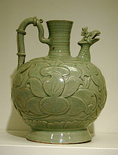 A green jug which is small at the bottom, wide near the middle, and has a tall, narrow opening at the top. The jug has a handle on one side and a carved bird head on the other side, bot connected to the top of the jug. The body of the jug has a flower or leaf pattern carved into it.