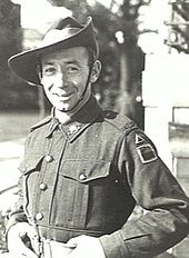 Black-and-white photo of a smiling, clean-shaven man wearing combat fatigues and a slouch hat. He is standing hands on hips with his hands gripping his light-coloured belt.