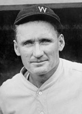 A shoulders-up picture of a smiling man in a white baseball uniform. He is wearing a dark-colored baseball cap on his head with a white block "W" on the front.
