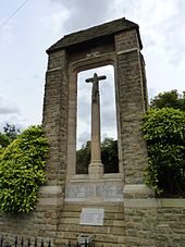 A photograph of a tall stone monument with a crucifix in the centre and a small v-shaped roof supported by two pillars. The names of those who fell in the two world wars are carved in stone at the base of the monument