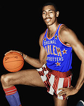 A black basketball player, wearing red and white pants and blue jersey and holding a basketball, stands with his right leg up
