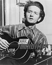 Half-length photo of a middle-aged man sitting in front of a closed door and playing a guitar and singing. His wavy black hair is partly covered  by a black hat tipped at a rakish angle. He wears a striped flannel work shirt. His black guitar has a sign on it that says, "This machine kills fascists".