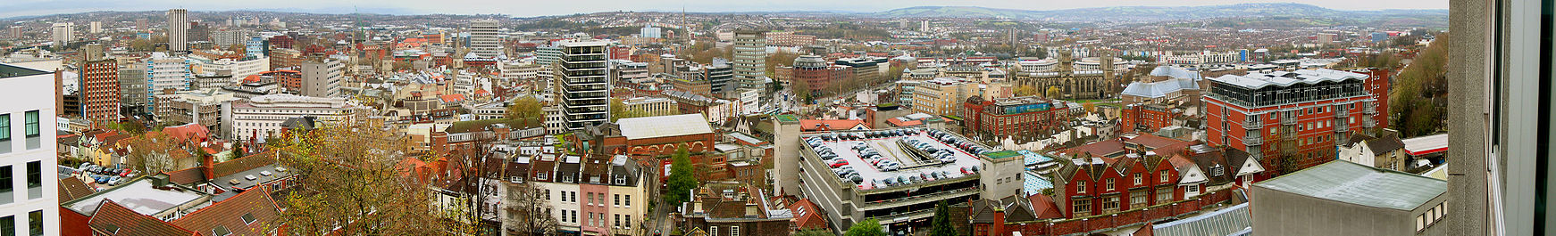A panoramic view looking over a cityscape of office blocks, old buildings, church spires and a multi-story car park. In the distance hills.