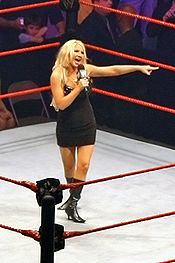 A blonde woman wearing a short black dress and black boots speaks into a microphone while standing in a wrestling ring with red ropes. She is pointing with her left hand.