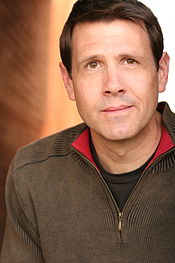 A brown-haired man wearing a brown fleece jacket looks directly ahead.