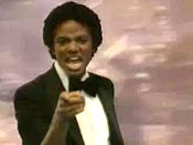 A adult African American male with short black hair. He is pointing forward with his right hand and is making a facial expression and he is wearing a black bow tie with a black and white tuxedo. Behind him, there is a pink background.