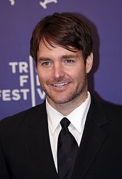 A smiling man with brown hair wearing a black suit jacket and black tie.