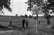 Two people stand either side of a lowered border pole on a dirt road with a sign in the foreground