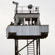 On top of a tubular steel support structure with a ladder there is a small cabin with windows to all sides, surrounded by a balcony on which stand two men in uniform, looking at the observer through binoculars; a third man is inside the cabin. There are searchlights on the tower's flat roof, and a railing around the roof's outer edge.