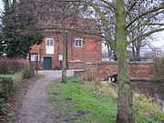 A view of the mill building from the footpath on the north bank.  The pool and the bridge across the race are visible to the right, but the red brick face of the building dominates. In the extreme foreground on the left is a bare-branched tree, its trunk lightly flecked with lichen. In the middle distance on the left is a holly, dark green and dense.
