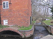 Close view of the south side of the building.  A small eliptical brick arch leads the water into the enclosed millrace, and there is an overflow weir to the right of the building. Trees are visible on the riverbank beyond.  The water is constrained in a red brick channel with a bulnose (curved) corner.  Two scoll shaped iron 'plates' are on the upper wall, terminals for tie-rods that pass through the building. This is a winter view, and the state of the trees makes this stark. There is a small and unturbulent flow of water through the overflow wier, because the level is accurately controlled by the main weir in the old lock chamber. A precarious plank bridge, with no handrails, crosses the overflow race to reach the lockside on the right.