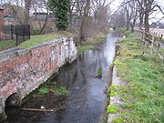 View downstream of the lock chamber build for the Sleaford Navigation, to maintain the head at the mill.  The chamber is largely brick built with stonde details for load-bearing parts, and the brick is coloured with moss and lichen.  A little desultory grass covers the top sides.  There are no lower gates, the lock having been converted into a wier many years ago.  A cheap iron railing fence, painted black recently, delineates the property associated with the mill and restaurant to the left. This is a winter view and many bare trees line the banks downstream. The trunks of the nearest can be clearly seen to be covered in ivy.  The water looks clear and placid.