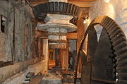 on the floor below the stones is the drive from the wheel.  We see the same vetical shaft that goes up through the floor, topped here with a black iron bevel gear.  Another bevel gear on the right, currently disengaged, is driven by the waterwheel. The room is cramped and whitewashed.