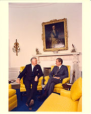 Two men sit in golden-yellow chairs and appear to be conversing cordially.